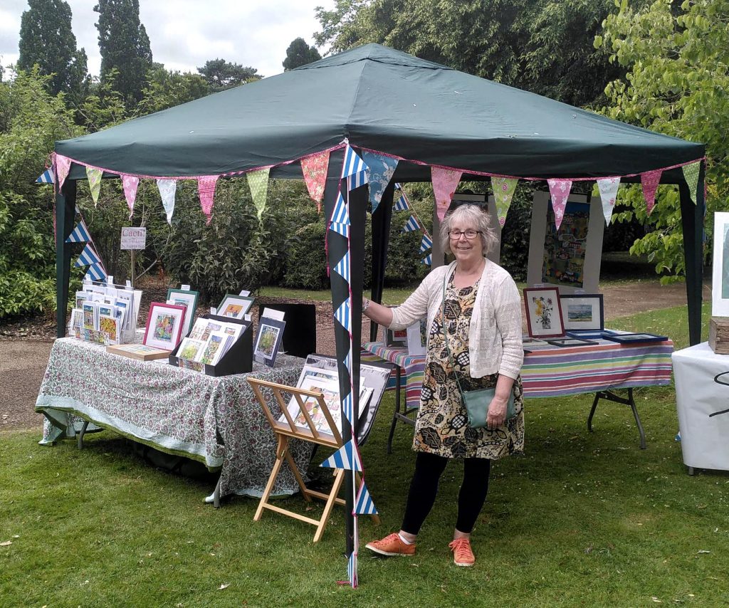 Helen standing next to a gazebo with a display of her work on tables in the grounds of the Leicester Botanic Garden.